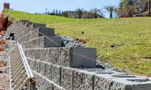 Budowa murek oporowy w ogrodzie. Betonowe bloki układane są warstwami, tworząc solidną konstrukcję. W tle zielona skarpa i dom.