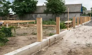 Solidny fundament pod ogrodzenie z drewnianymi słupkami i betonowym murem. W tle widać budowę domu.