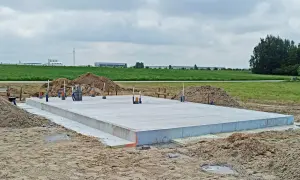 Płyta fundamentowa koszt budowy. Betonowa płyta fundamentowa z wystającymi rurami i przewodami, otoczona piaskiem i ziemią. W tle zielone pola i budynki przemysłowe.