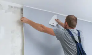 Mężczyzna tapetuje ścianę, wygładzając papier narzędziem. Sprawdź, jaka jest tapetowanie cena za metr kwadratowy.