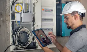 Technik w kasku i okularach ochronnych wykonuje przegląd instalacji elektrycznej, korzystając z tabletu z aplikacją diagnostyczną.