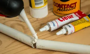 Aplikator z klejem naprawia pękniętą białą rurkę. Obok leżą tubki kleju, idealne, by dowiedzieć się, czym skleić plastik.