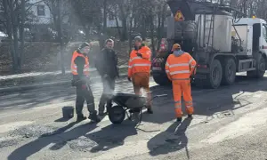 Ekipa drogowców w pomarańczowych strojach pracuje przy drodze, obok wózka i specjalistycznej ciężarówki.