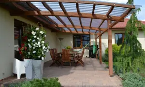 Drewniana pergola z przezroczystym dachem nad tarasem, gdzie stoi stół z krzesłami, grill i hamak. Idealne miejsce, co na taras.