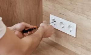Ręce majsterkowicza dokręcają śrubki w białej ramce z trzema gniazdkami. Widać drewnianą ścianę, a w gniazdku jest napięcie.