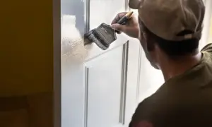 Mężczyzna w czapce z daszkiem maluje białe drzwi pędzlem. Odświeża wygląd wnętrza.