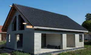 Dom w stanie surowym otwartym, z dachem pokrytym czarną dachówką. Widoczne ściany z bloczków, otwory okienne i drzwiowe.