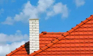 Pomarańczowy dach wielospadowy z czerwonej dachówki i biały komin na tle błękitnego nieba z chmurami.