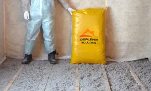 Osoba w kombinezonie ochronnym stoi obok worka z materiałem do ocieplenia celulozą. Podłoga pokryta jest warstwą izolacji.