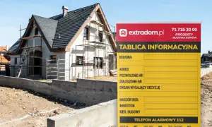 Tablica informacyjna budowy domu w trakcie prac, z widocznym rusztowaniem i fundamentami. Na tablicy dane inwestora i numer telefonu alarmowego.