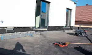 Prace na dachu płaskim: drabina, rolka do papy i czarny worek z odpadami.