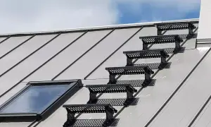 Sześć czarnych stopni bezpieczeństwa na metalowym dachu, prowadzących do okna dachowego. Solidne opierzenie dachu zapewnia bezpieczne przejście.