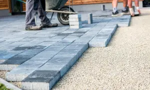 Układanie płyt betonowych na podjeździe – alternatywa dla kostki. Pracownicy budowlani układają szare płyty na podsypce żwirowej.