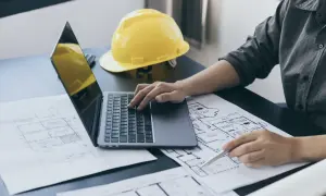 Inżynier przy laptopie analizuje plany, obliczając powierzchnię całkowitą budynku. Obok leży żółty kask.