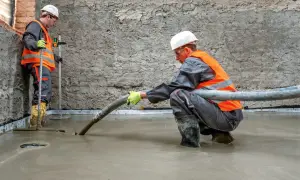 Dwóch budowlańców w kamizelkach odblaskowych wylewa pianobeton przez wąż na podłogę.
