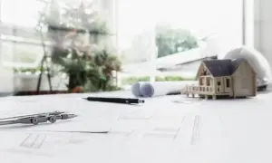 Model domu, cyrkiel i plany architektoniczne na stole. W tle okno z zielenią. Projekt uwzględnia nieprzekraczalna linia zabudowy.