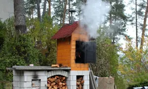 Dym unosi się z drewnianej wędzarni ogrodowej, pokazując, jak zbudować wędzarnię. Widać stosy drewna i wiszące kiełbaski.
