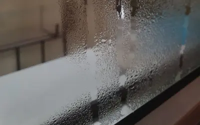 Zaparowane okna po nocy, z kroplami wody tworzącymi wzory na szybie. W tle widać niewyraźne kontury mebli i jasne światło.