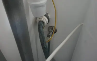Szara rura odpływowa pralki podłączona do syfonu. Widoczny żółty przewód uziemiający i biały kabel.