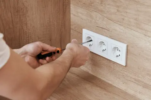 Ręce majsterkowicza dokręcają śrubki w białej ramce z trzema gniazdkami. Widać drewnianą ścianę, a w gniazdku jest napięcie.