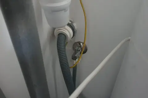 Szara rura odpływowa pralki podłączona do syfonu. Widoczny żółty przewód uziemiający i biały kabel.