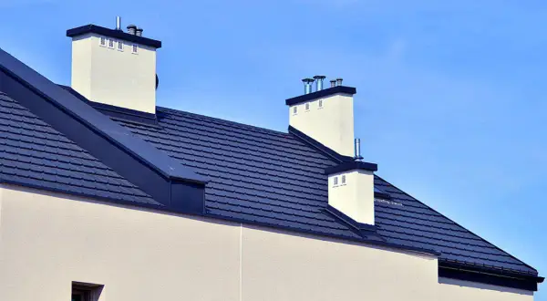 Nowoczesny dach bezokapowy z ciemnogranatową dachówką i białymi kominami, pod błękitnym niebem.