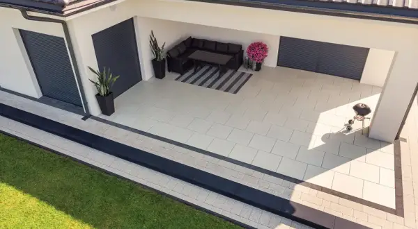 Nowy, nowoczesny chodnik z płyt betonowych otacza dom. Na tarasie wygodna sofa i grill czekają na gości.