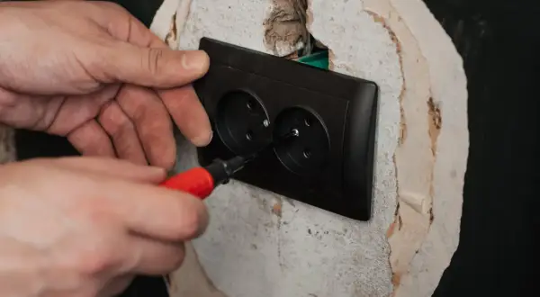 Ręce montują czarne podwójne gniazdko elektryczne. Śrubokręt z czerwoną rączką dokręca śrubę, kończąc podłączenie gniazdka.