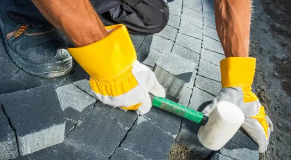 Pracownik w żółtych rękawicach układa kostkę brukową na betonie, używając gumowego młotka.