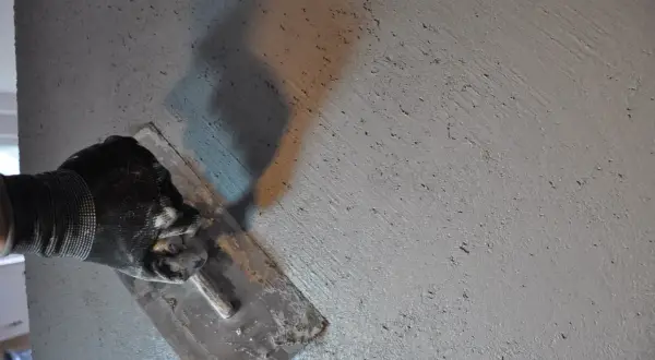 Ręka w rękawicy wygładza świeżą zaprawę, pokazując, jak zrobić beton. Cień narzędzia rzuca się na chropowatą powierzchnię.