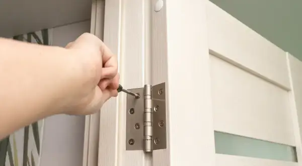 Dłoń dokręca śrubę w zawiasie drzwi, wykonując regulację zawiasów drzwi.