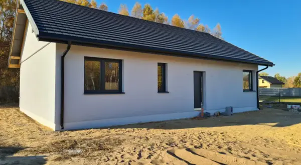 Nowy dom w stanie surowym zamkniętym, z szarym dachem i białymi ścianami, otoczony piaskiem i jesiennymi drzewami.