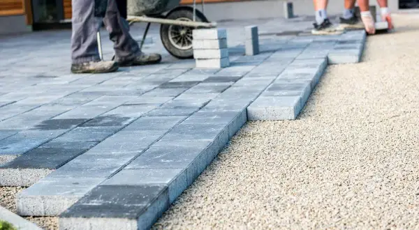 Układanie płyt betonowych na podjeździe – alternatywa dla kostki. Pracownicy budowlani układają szare płyty na podsypce żwirowej.