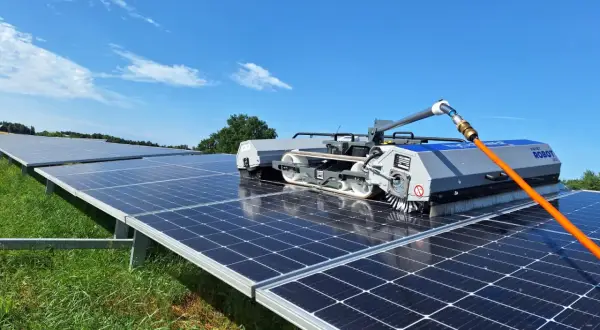 Robot do mycia paneli fotowoltaicznych czyści rzędy paneli na zielonej łące pod błękitnym niebem.