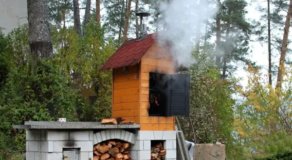 Dym unosi się z drewnianej wędzarni ogrodowej, pokazując, jak zbudować wędzarnię. Widać stosy drewna i wiszące kiełbaski.
