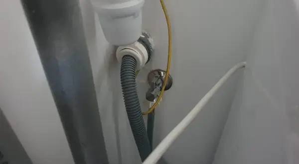 Szara rura odpływowa pralki podłączona do syfonu. Widoczny żółty przewód uziemiający i biały kabel.