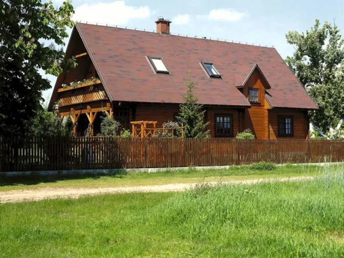 Drewniana zabudowa zagrodowa z werandą i drewnianym płotem, otoczona zielenią.