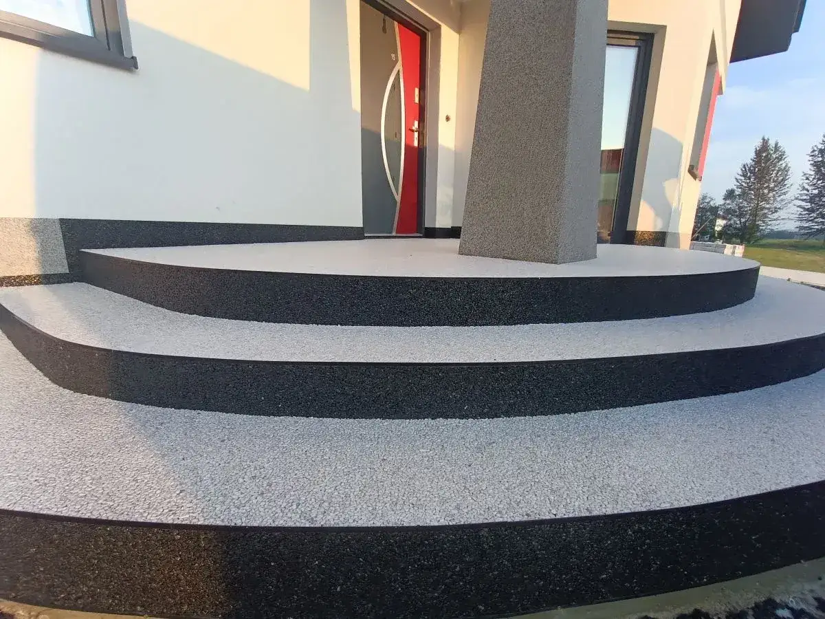 Nowe schody z kamiennym dywanem, koszt 1m2 to inwestycja w nowoczesny wygląd domu. Jasny gres i czarne obrzeża tworzą elegancką całość.