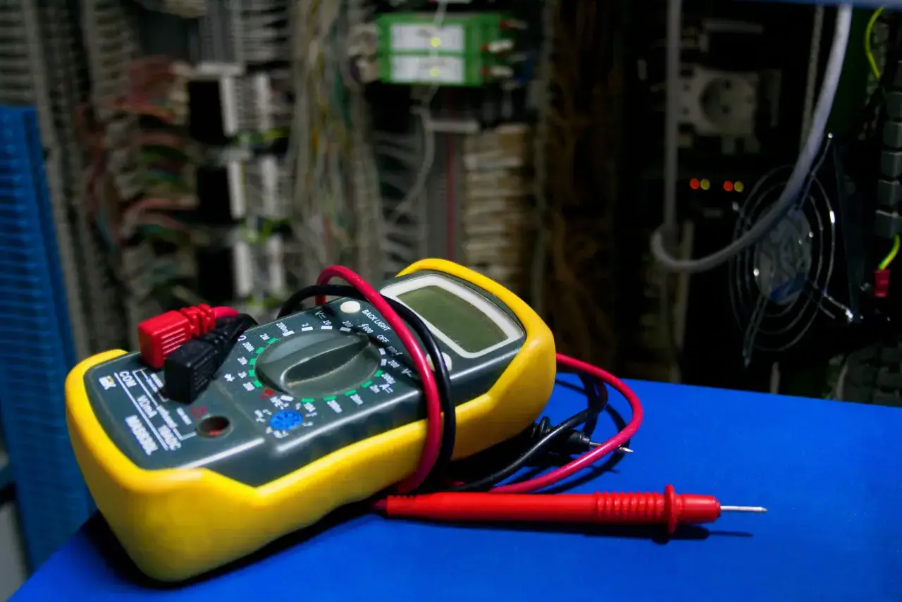 Żółty galwanometr z czerwonymi i czarnymi przewodami leży na niebieskiej powierzchni, w tle widać skomplikowaną instalację elektryczną.