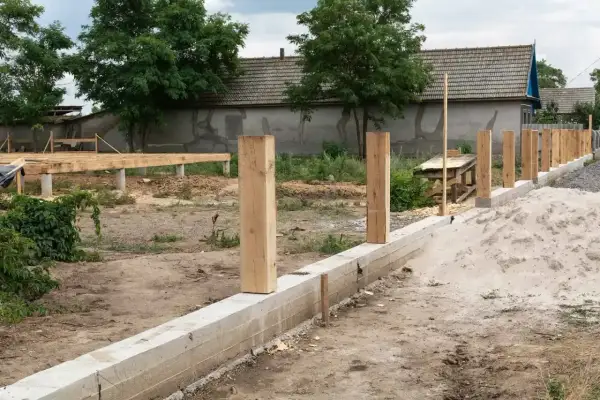 Solidny fundament pod ogrodzenie z drewnianymi słupkami i betonowym murem. W tle widać budowę domu.