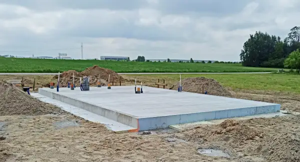 Płyta fundamentowa koszt budowy. Betonowa płyta fundamentowa z wystającymi rurami i przewodami, otoczona piaskiem i ziemią. W tle zielone pola i budynki przemysłowe.