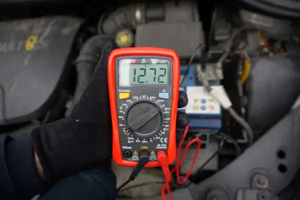 Ręka w czarnej rękawiczce trzyma multimetr pokazujący 12.72V. To minimalne napięcie akumulatora do uruchomienia.