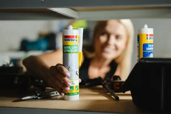 Kobieta trzyma tubkę akrylu lekkiego szpachlowego Soudal. Akryl to wszechstronny materiał budowlany.