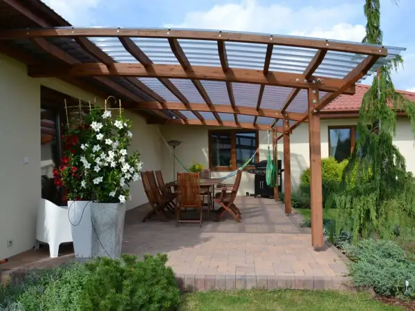 Drewniana pergola z przezroczystym dachem nad tarasem, gdzie stoi stół z krzesłami, grill i hamak. Idealne miejsce, co na taras.