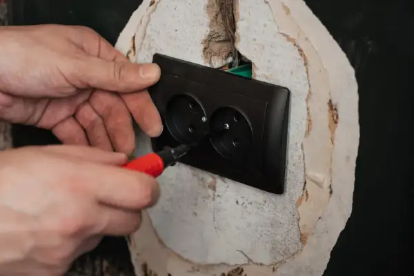 Ręce montują czarne podwójne gniazdko elektryczne. Śrubokręt z czerwoną rączką dokręca śrubę, kończąc podłączenie gniazdka.