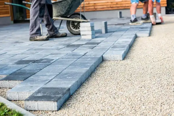 Układanie płyt betonowych na podjeździe – alternatywa dla kostki. Pracownicy budowlani układają szare płyty na podsypce żwirowej.