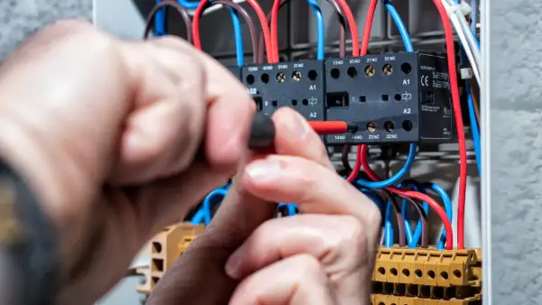 Ręce majsterkowicza przykręcają śrubę w skrzynce elektrycznej, gdzie widać przewody i styczniki. Kontrola napięcia prądu.