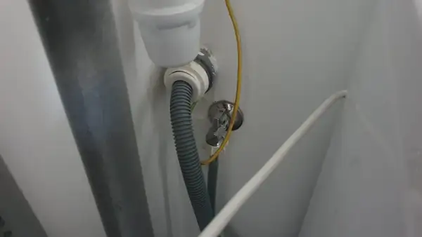 Szara rura odpływowa pralki podłączona do syfonu. Widoczny żółty przewód uziemiający i biały kabel.